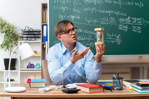 Jonge mannelijke leraar die een bril draagt met een zandloper die les voorbereidt en er verward uitziet terwijl hij aan de schoolbank zit met boeken en notities voor het bord in de klas