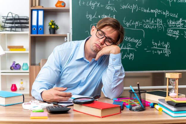 Jonge mannelijke leraar die een bril draagt, moe en verveeld, wil slapen aan de schoolbank met boeken en notities voor het bord in de klas