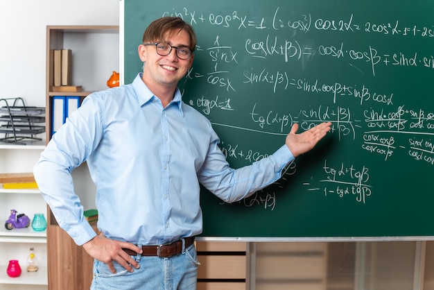 Jonge mannelijke leraar met een bril die de les gelukkig en positief glimlachend uitlegt terwijl hij in de buurt van het bord staat met wiskundige formules in de klas