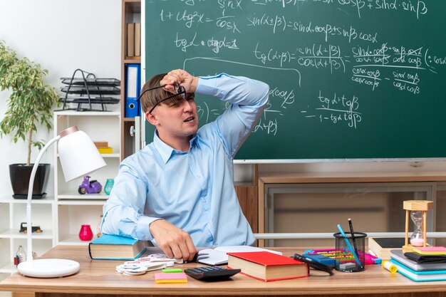 Jonge mannelijke leraar met een bril die er verward en erg angstig uitziet terwijl hij aan de schoolbank zit met boeken en notities voor het bord in de klas