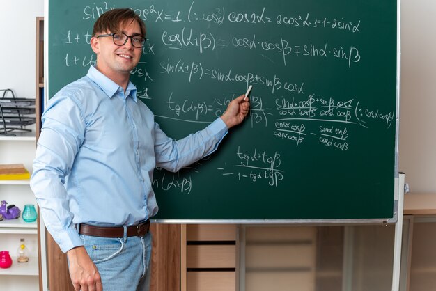 Jonge mannelijke leraar met een bril die krijt vasthoudt en les uitlegt die zelfverzekerd glimlacht en in de buurt van het bord staat met wiskundige formules in de klas