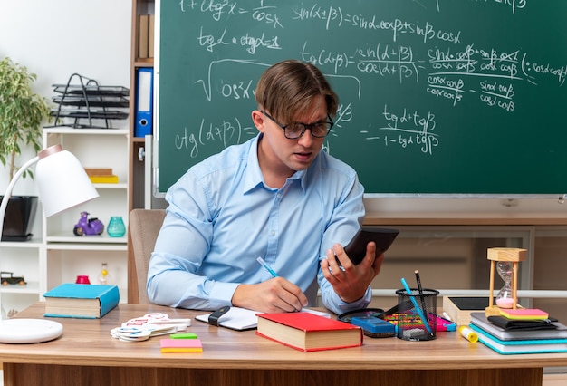 Jonge mannelijke leraar met een bril die naar zijn smartphone kijkt en er zelfverzekerd uitziet terwijl hij aan de schoolbank zit met boeken en notities voor het bord in de klas