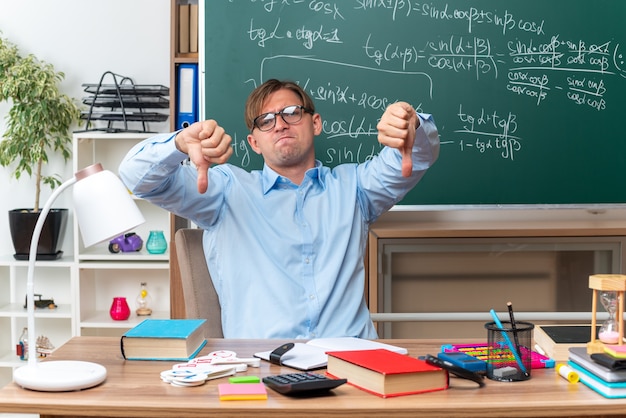 Jonge mannelijke leraar met een bril die ontevreden is met duimen naar beneden zittend aan de schoolbank met boeken en notities voor het bord in de klas