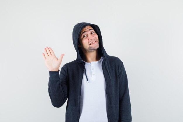 Jonge mannelijke zwaaiende hand om afscheid te nemen in t-shirt, jas en op zoek vrolijk, vooraanzicht.