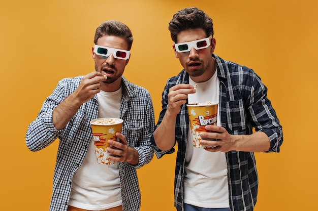 Jonge mannen in geruite overhemden en witte t-shirts kijken naar film in 3D-brillen en eten popcorn op een oranje geïsoleerde achtergrond