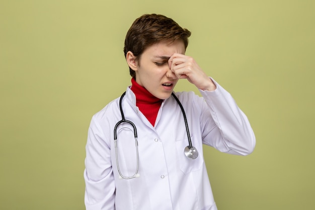 Jonge meisjesdokter in witte jas met stethoscoop om nek die er onwel moe en onwerkt uitziet en nerneus aanraakt tussen gesloten ogen die op groen staan