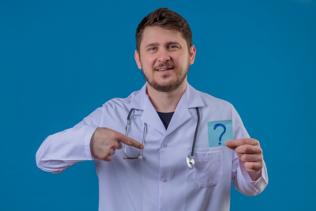Jonge mens arts die witte laag en stethoscoopholdingsdocument met vraagteken met glimlach op gezicht dragen die vinger aan zich over geïsoleerde blauwe achtergrond richten