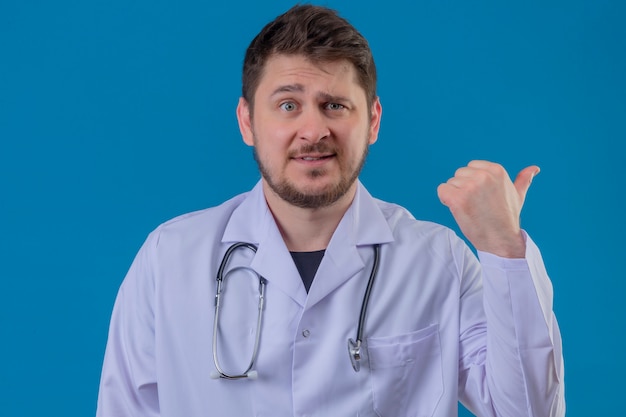 Jonge mens arts witte laag dragen en stethoscoop die aan de rug erachter met hand en duimen over geïsoleerde blauwe achtergrond richten