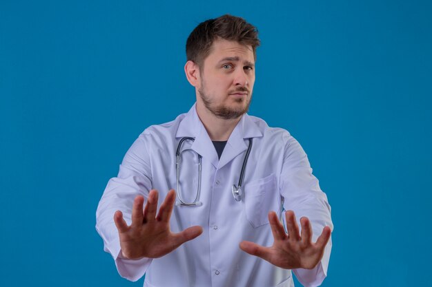 Jonge mens arts witte laag dragen en stethoscoop die eindeteken, verdedigingsgebaar doen over geïsoleerde blauwe achtergrond