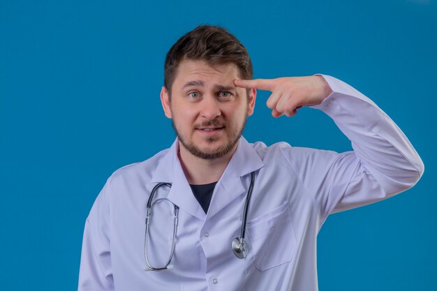 Jonge mens arts witte laag dragen en stethoscoop die verward wijzend vinger aan zijn hoofd over geïsoleerde blauwe achtergrond kijken