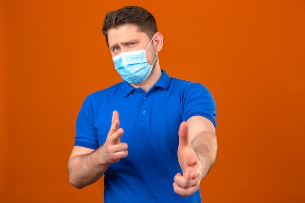 Jonge mens die blauw poloshirt in medisch beschermend masker draagt dat met vingers aan camera met sceptische uitdrukking richt die zich over geïsoleerde oranje muur bevindt