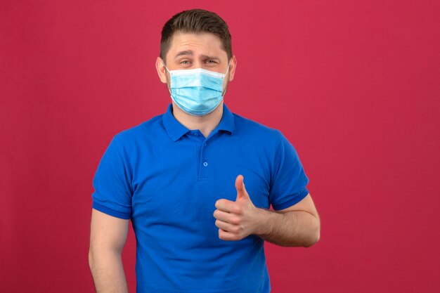 Jonge mens die blauw poloshirt in medisch beschermend masker draagt dat vrolijk tonend duim over geïsoleerde roze muur glimlacht