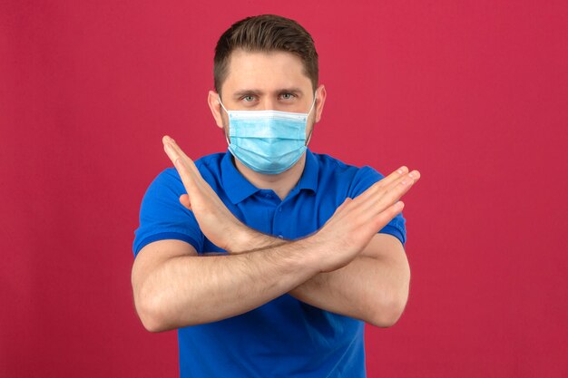 Jonge mens die blauw poloshirt in medisch beschermend masker draagt dat zich met gekruiste wapens bevindt die eindegebaar met fronsend gezicht over geïsoleerde roze muur maakt