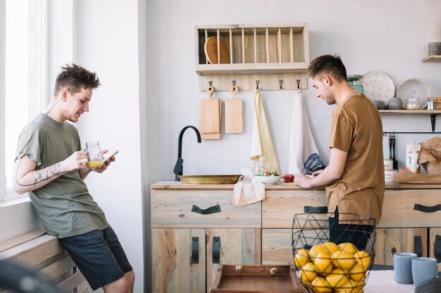 Jonge mens die cellphone gebruiken terwijl zijn vriend die voedsel in keuken voorbereidt