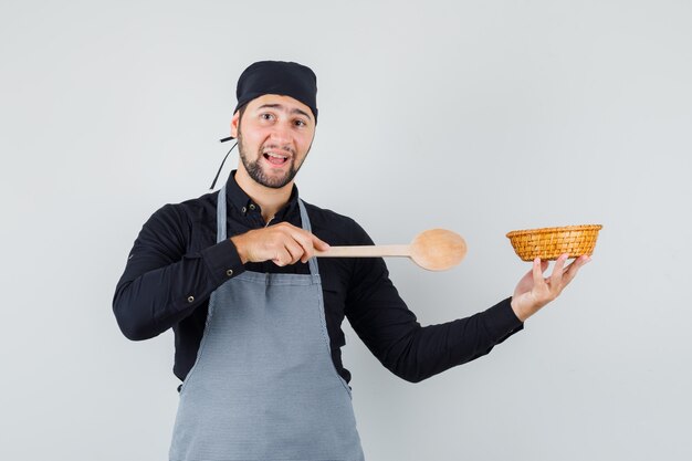 Jonge mens die houten lepel richt op rieten mand in overhemd, schort en vrolijk kijkt. vooraanzicht.