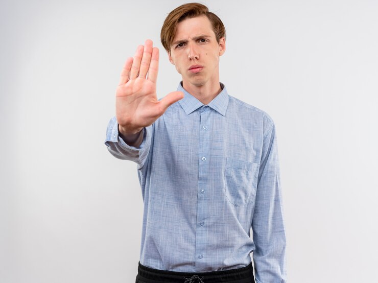 Gratis Foto jonge mens die in blauw overhemd stopteken met open hand met ernstig gezicht maakt dat zich over witte muur bevindt