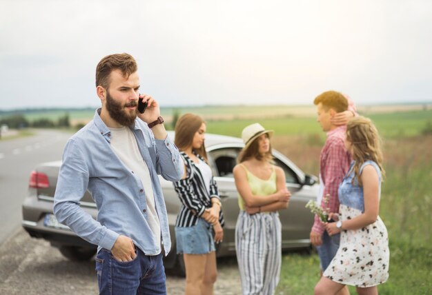 Gratis foto jonge mens die op mobiele telefoon spreekt die zich voor vrienden bij wegreis bevindt