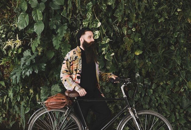 Gratis foto jonge mens die zich met fiets voor groene installatiesmuur bevindt