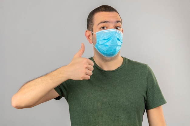 Jonge mens in medisch masker die ok teken, duim omhoog met vingers doen die op wit worden geïsoleerd