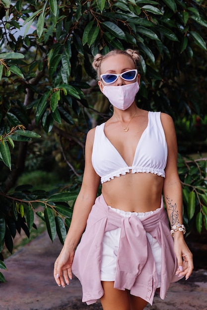 Jonge mode vrouw in witte crop top, zonnebril en wit medisch gezichtsmasker in de natuur