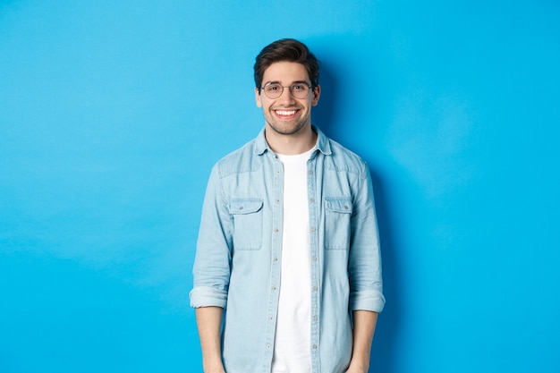 Jonge moderne man met een bril en een casual outfit die tegen een blauwe achtergrond staat, gelukkig glimlachend in de camera