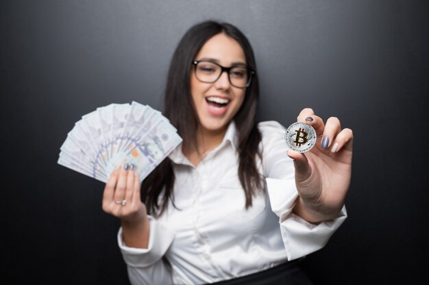 Jonge moderne succesvolle bedrijfsvrouw die een gouden bitcoin en contant gelddollar houdt die op zwarte muur wordt geïsoleerd