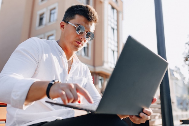 Jonge modieuze kerel in overhemd met telefoon en notitieboekje op bank op zonnige warme freelance dag in openlucht