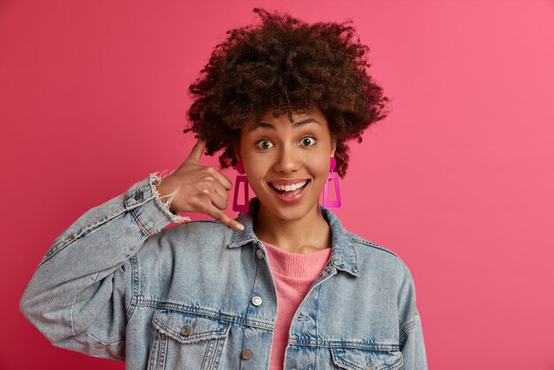 Jonge modieuze vrouw maakt telefoongebaar, vraagt om haar te bellen, drukt positieve emoties uit, draagt denim kleding, glimlacht gelukkig, geïsoleerd op roze muur. Bel me terug teken. Lichaamstaal