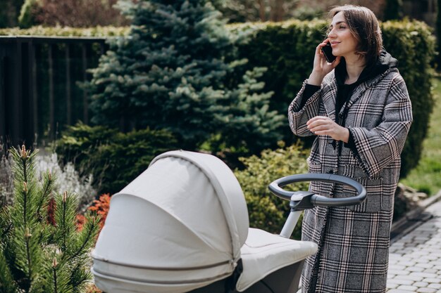 Jonge moeder die met kinderwagen in park loopt en op de telefoon spreekt