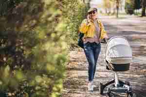 Gratis foto jonge moeder die met kinderwagen in park loopt en telefoon met behulp van