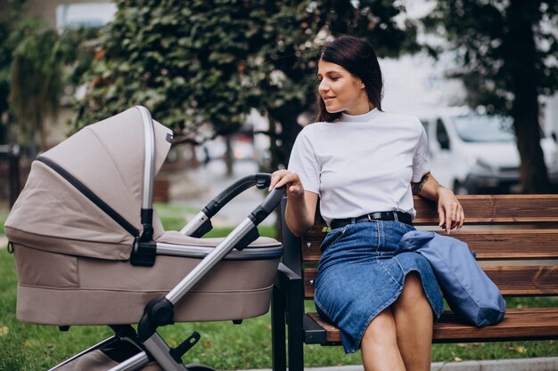 Jonge moeder zittend op een bankje in park met kinderwagen