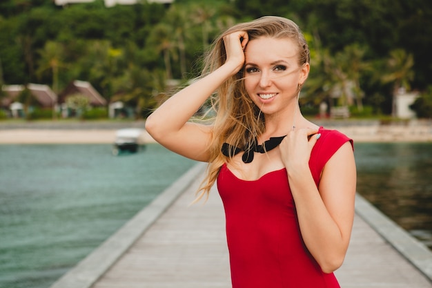 Jonge mooie aantrekkelijke vrouw die alleen staat op de pier in een luxe resorthotel, zomervakantie, rode lange jurk, blond haar, sexy kleding, tropisch strand, verleidelijk, sensueel, glimlachen