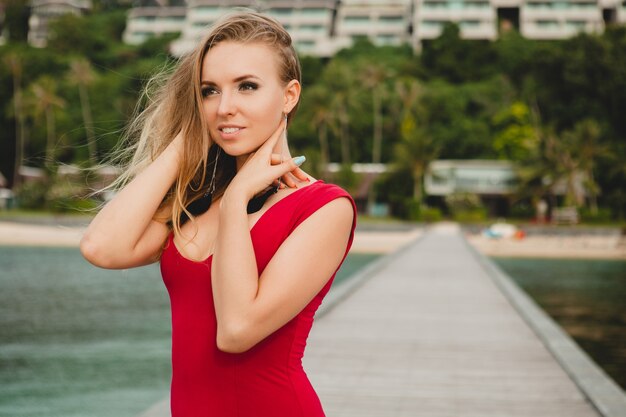 Jonge mooie aantrekkelijke vrouw die alleen staat op de pier in een luxe resorthotel, zomervakantie, rode lange jurk, blond haar, sexy kleding, tropisch strand, verleidelijk, sensueel, glimlachen