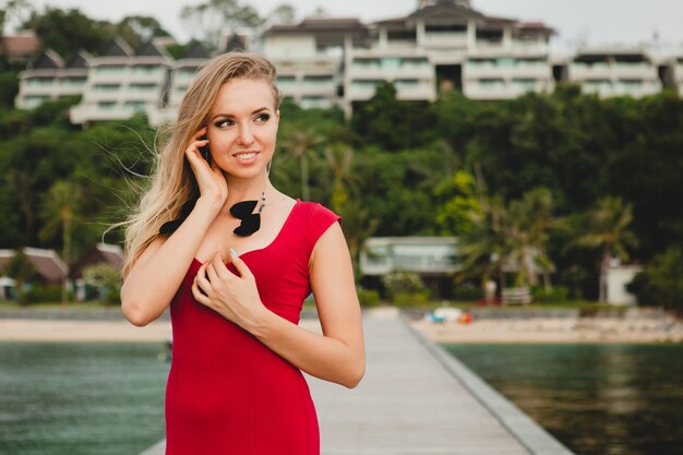 Jonge mooie aantrekkelijke vrouw die alleen staat op de pier in een luxe resorthotel, zomervakantie, rode lange jurk, blond haar, sexy kleding, tropisch strand, verleidelijk, sensueel, glimlachen