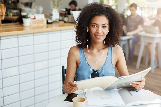 Jonge mooie Afrikaanse studente die in hoofdtelefoons in koffie het glimlachen holdingsboek zitten. Onderwijs en leren.