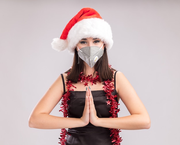 Jonge mooie blanke meisje dragen kerstmuts en beschermende masker klatergoud slinger rond nek kijken camera handen bij elkaar houden geïsoleerd op witte achtergrond