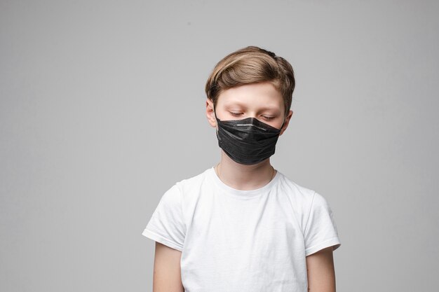 Jonge mooie blanke tiener in wit t-shirt, zwarte spijkerbroek staat met zwarte medische masker neerkijkt