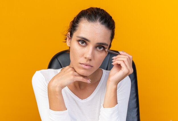 Jonge, mooie blanke vrouw die aan tafel zit met make-uptools die eyeliner toepassen die op een oranje muur is geïsoleerd met kopieerruimte