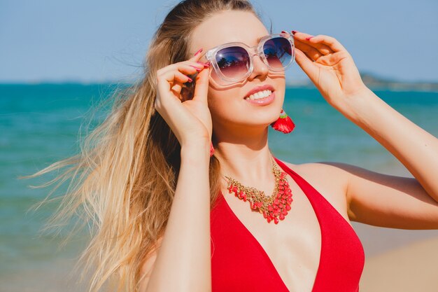 Jonge mooie blonde vrouw die op strand in rood zwemmend kostuum, zonnebril zonnebaden