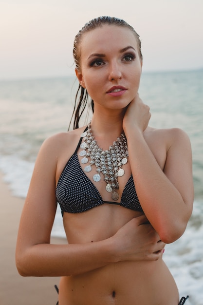 Jonge mooie blonde vrouw die op zandstrand in bikini zwemmend kostuum, uitstekende halsband zonnebaden