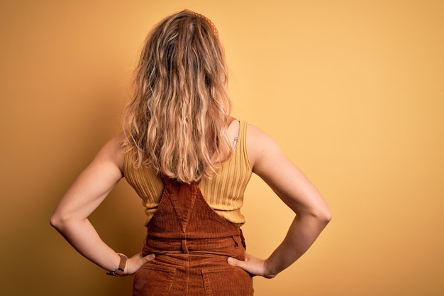 Jonge, mooie blonde vrouw, gekleed in een overall en diadeem die over een gele achtergrond staat en achteruit kijkt en wegkijkt met de armen op het lichaam