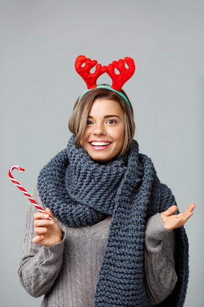 Gratis foto jonge mooie blonde vrouw in grote gebreide sjaal en kerst rendieren gewei glimlachend eten gestreepte lolly op grijs.