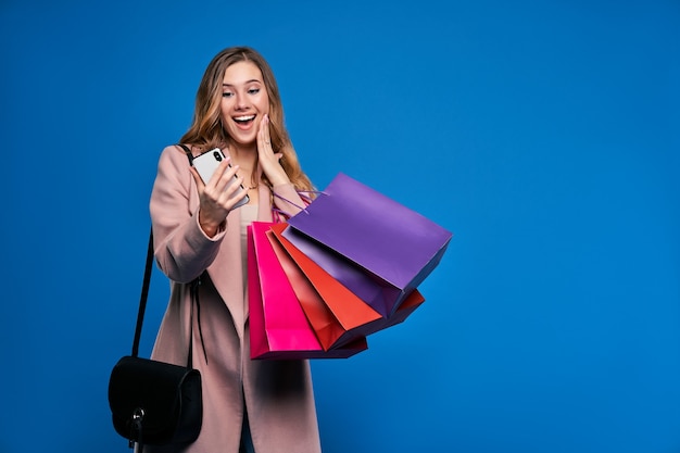 Jonge mooie blonde vrouw in jas op een blauwe muur met mobiele telefoon die online winkelen doet.