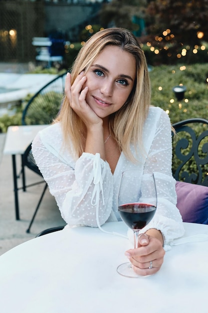 Jonge, mooie blonde vrouw in witte jurk die dromerig in de camera kijkt met een glas rode wijn in de tuin van restaurant