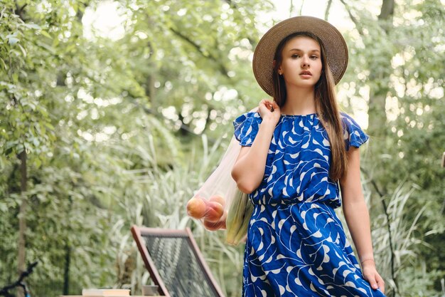 Jonge mooie bruinharige vrouw in blauwe jurk en hoed met eco-tassen met groenten en fruit op de schouder terwijl ze bedachtzaam in de camera in het park kijkt