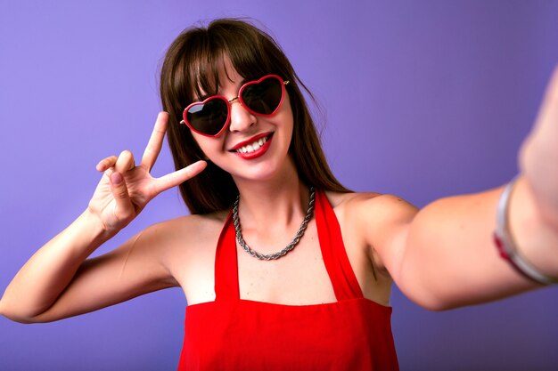 Jonge mooie brunette vrouw met lange haren en geweldige glimlach selfie maken op paarse achtergrond, vintage getinte kleuren, retro elegante outfit en hart zonnebril.