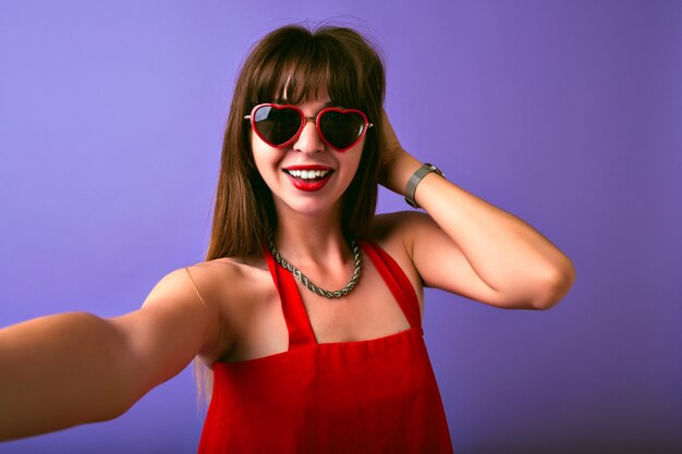 Jonge mooie brunette vrouw met lange haren en geweldige glimlach selfie maken op paarse achtergrond, vintage getinte kleuren, retro elegante outfit en hart zonnebril.