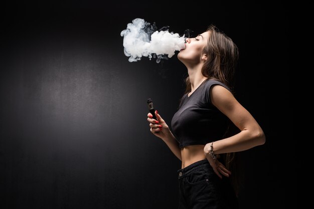 Jonge mooie brunette vrouw roken, vaping e-sigaret met rook op de zwarte muur