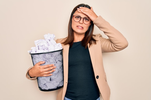 Jonge, mooie brunette zakenvrouw die een volle papierbak met gekreukt papier vasthoudt, gestrest en gefrustreerd met de hand op het hoofd, verrast en boos gezicht