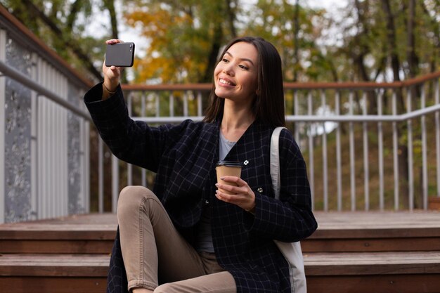 Jonge mooie casual vrouw met koffie om graag selfie te nemen op mobiel in herfstpark